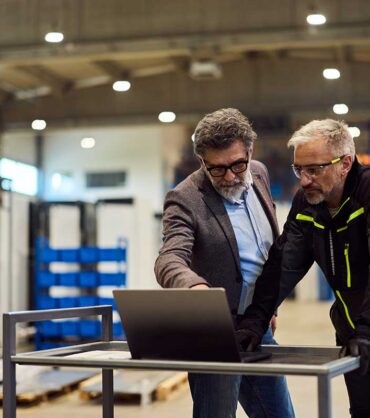 two-professionals-in-a-warehouse-looking-at-a-laptop two-professionals-in-a-warehouse-looking-at-a-laptop