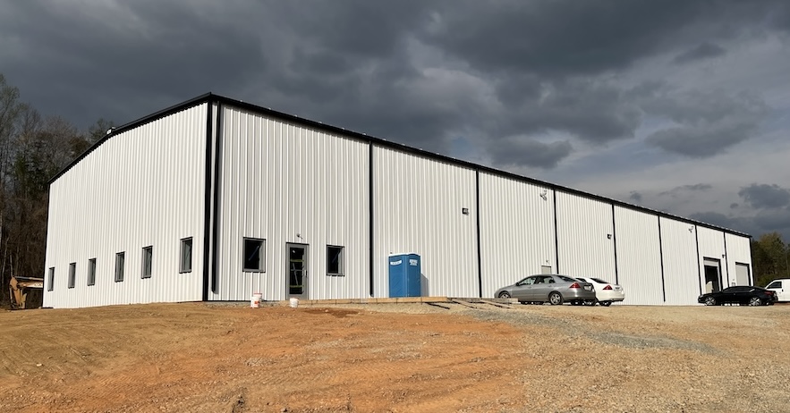 Large light-gray metal industrial warehouse with small windows along the side, blue portable toilets outside, and several cars parked on a gravel lot under a cloudy sky.